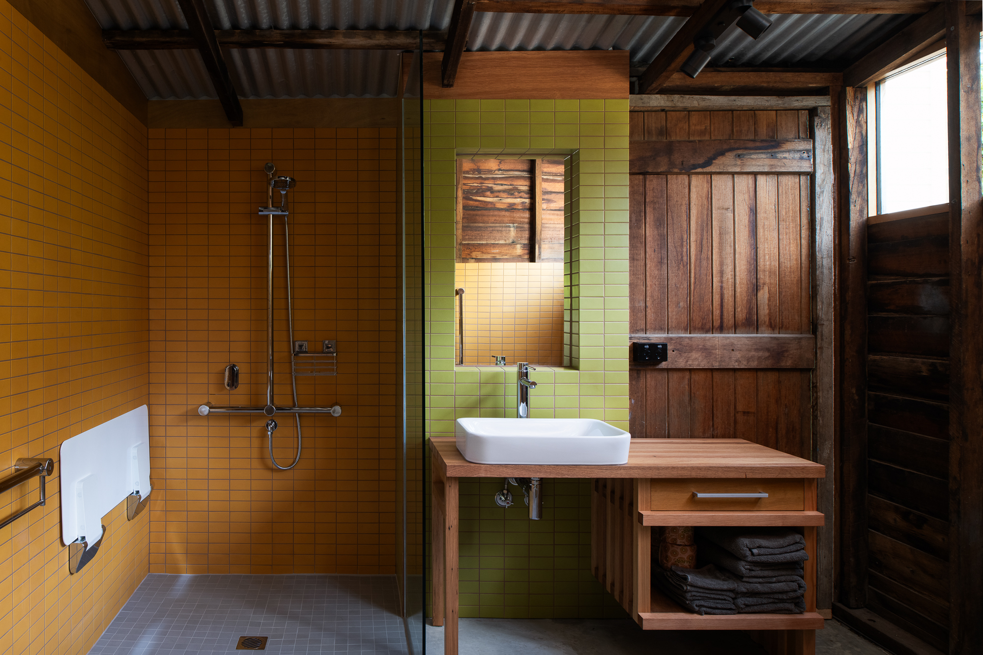 yellow tiled shower with white shower seat and green tiled mirror above basin on timber table.