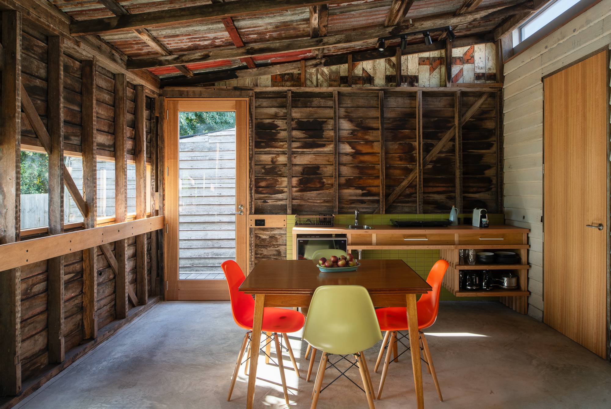 Room with timber walls and exposed framing. Midcentury modern dining table and chairs in front of a kitchenette.