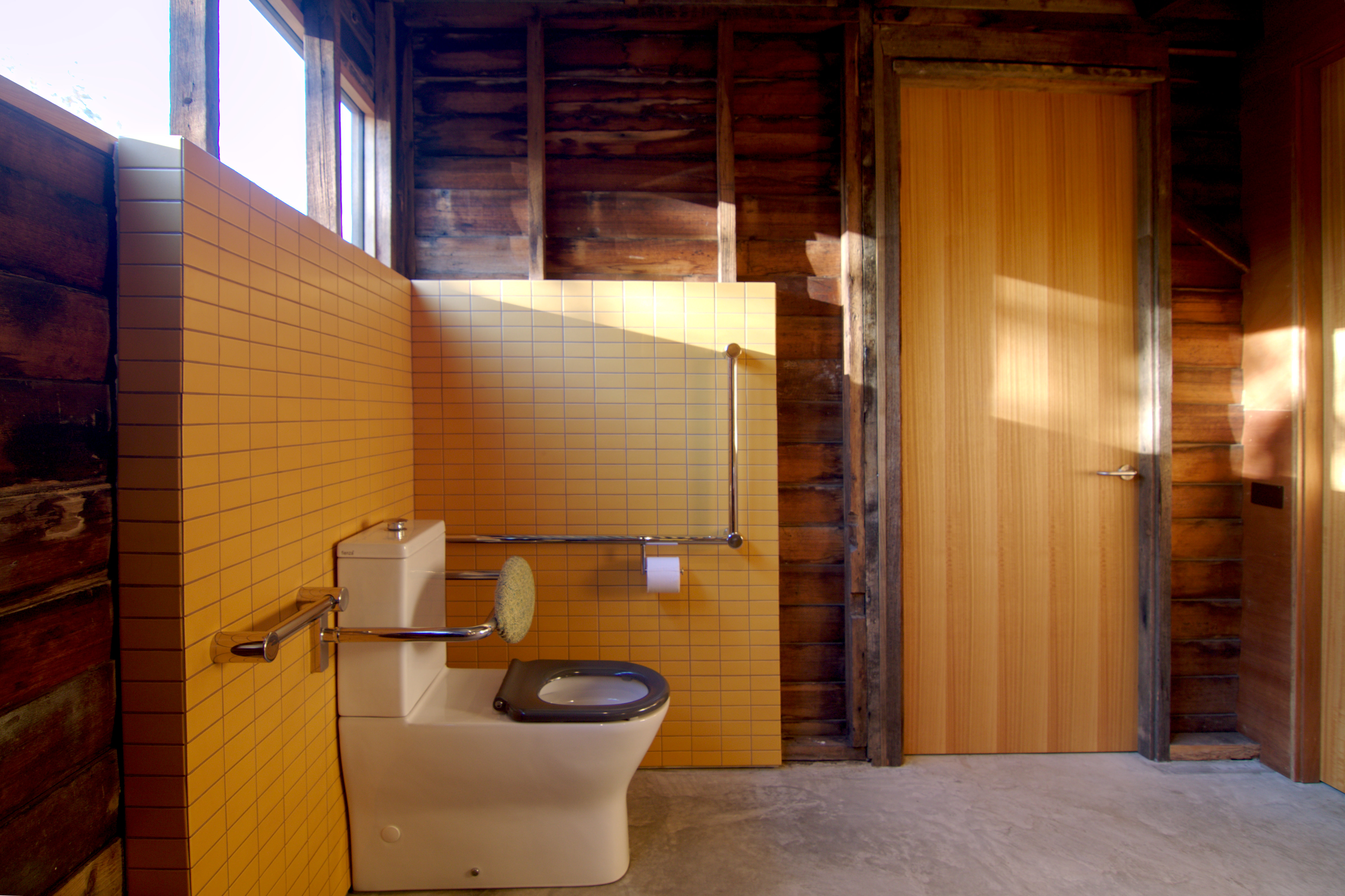 high window over white toilet with grey seat and green backrest with grabraiuls on yellow tiled wall