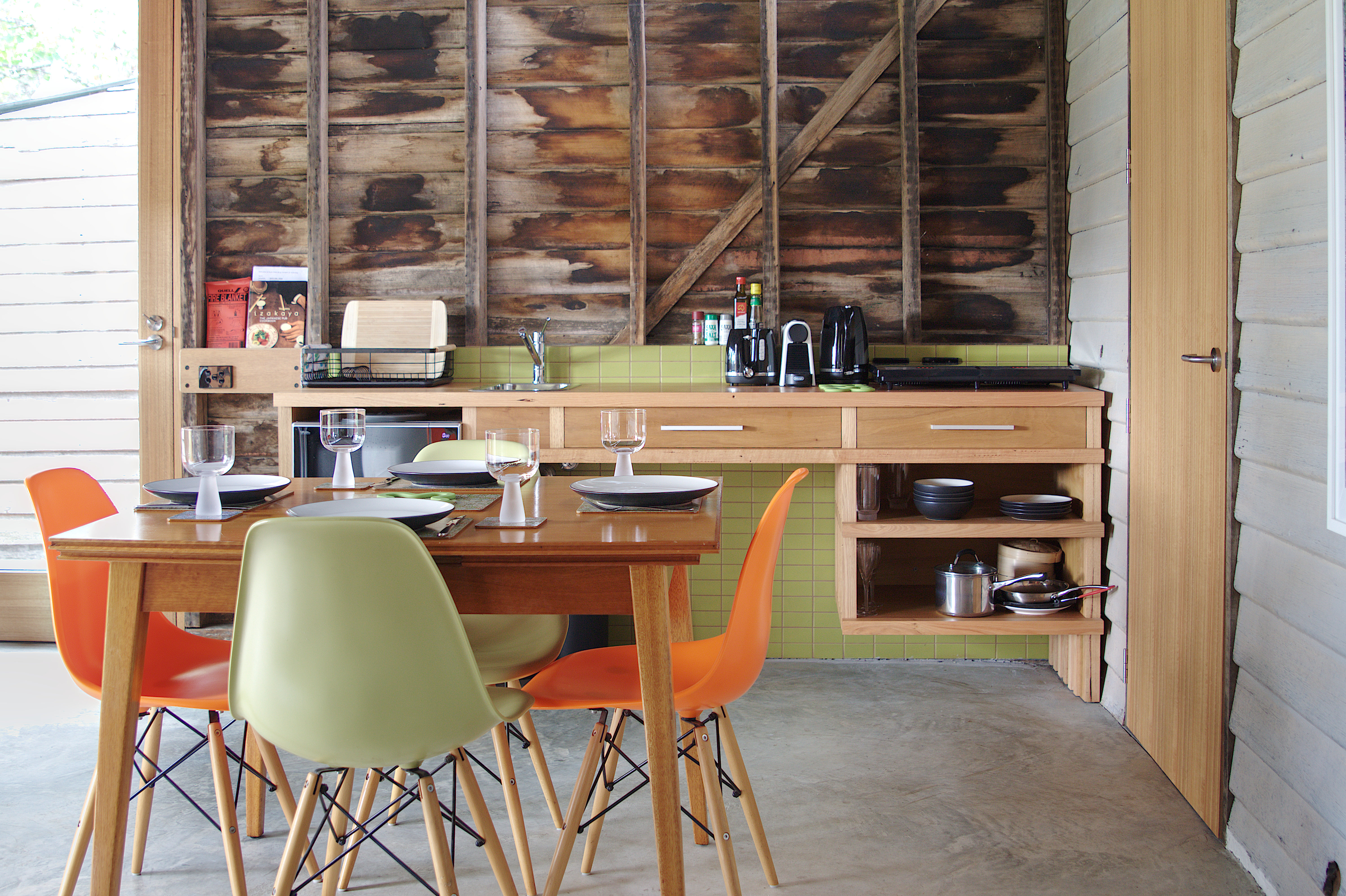 Mid century modern dining table and chairs in foreground. Timber kitchen bench in background with drawers, open shelves and green tile splashback. Timber walls and concrete floor.