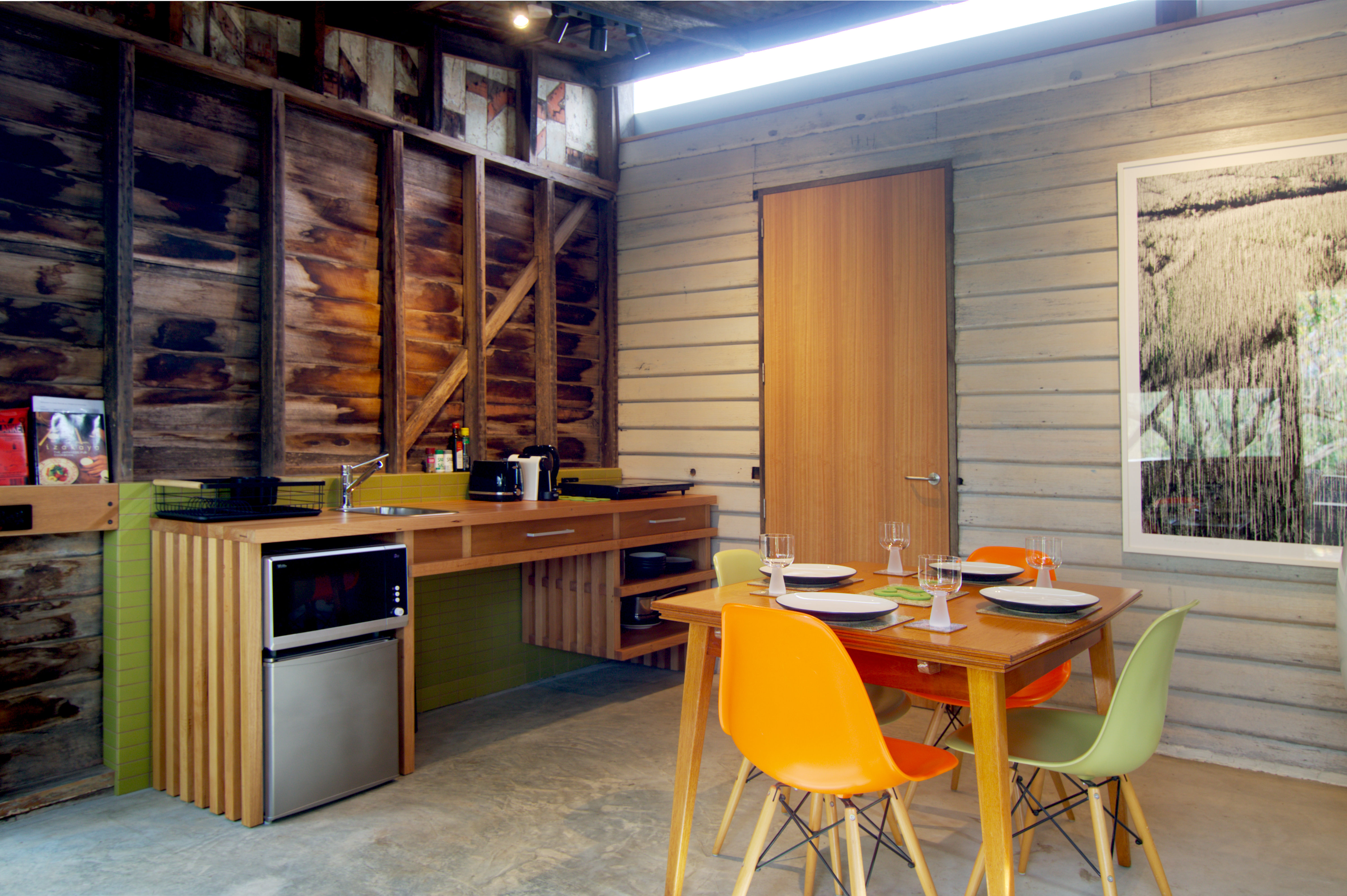 Highlight strip window above a door and artwork. Mid century modern dining table with plates and glasses and coloured chairs.  Kitchen bench open underneath with microwave and bar fridge at end.