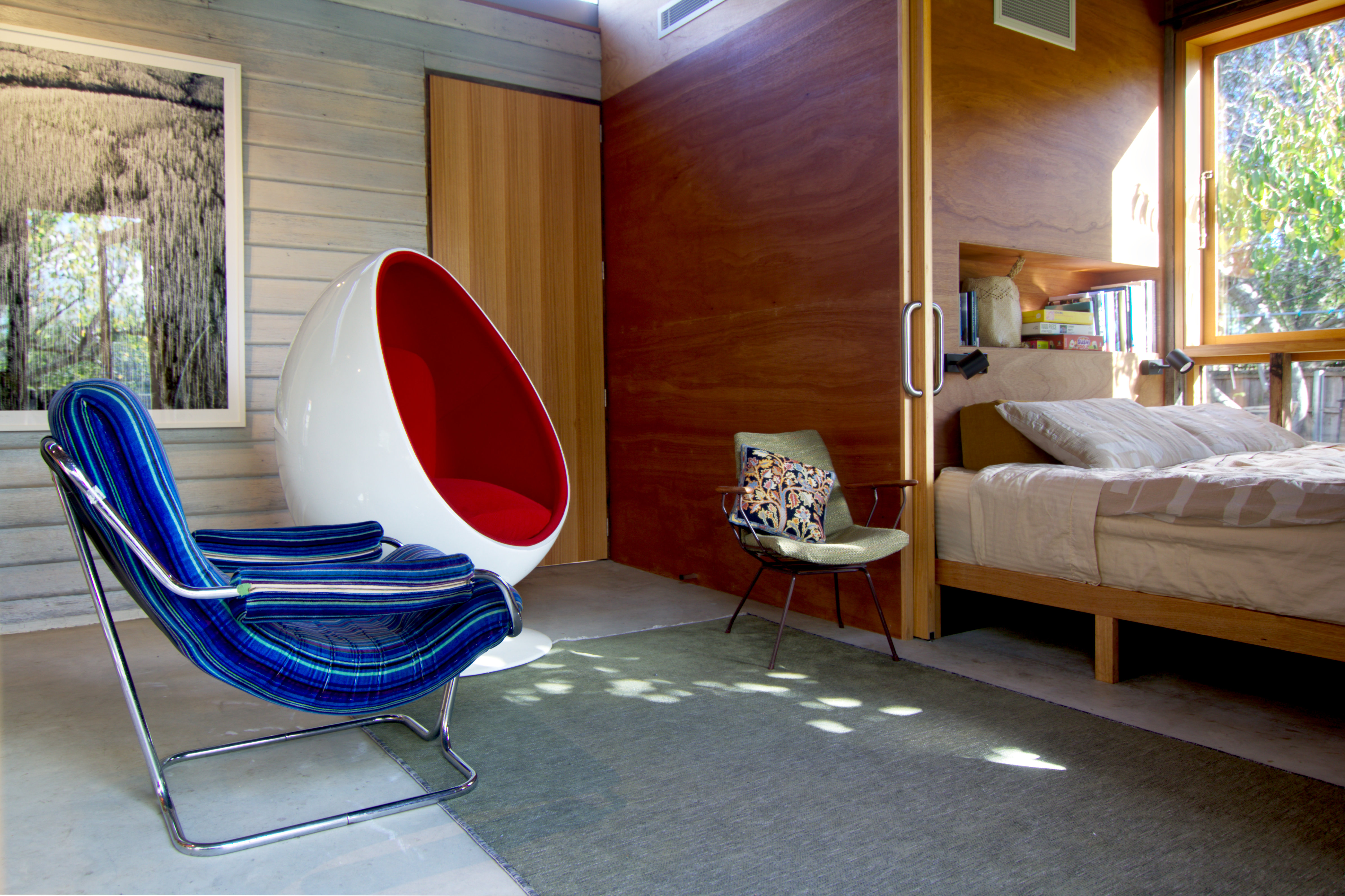 blue sling chair, red and white egg chair and green chair in front of timber wall with day bed in alcove.