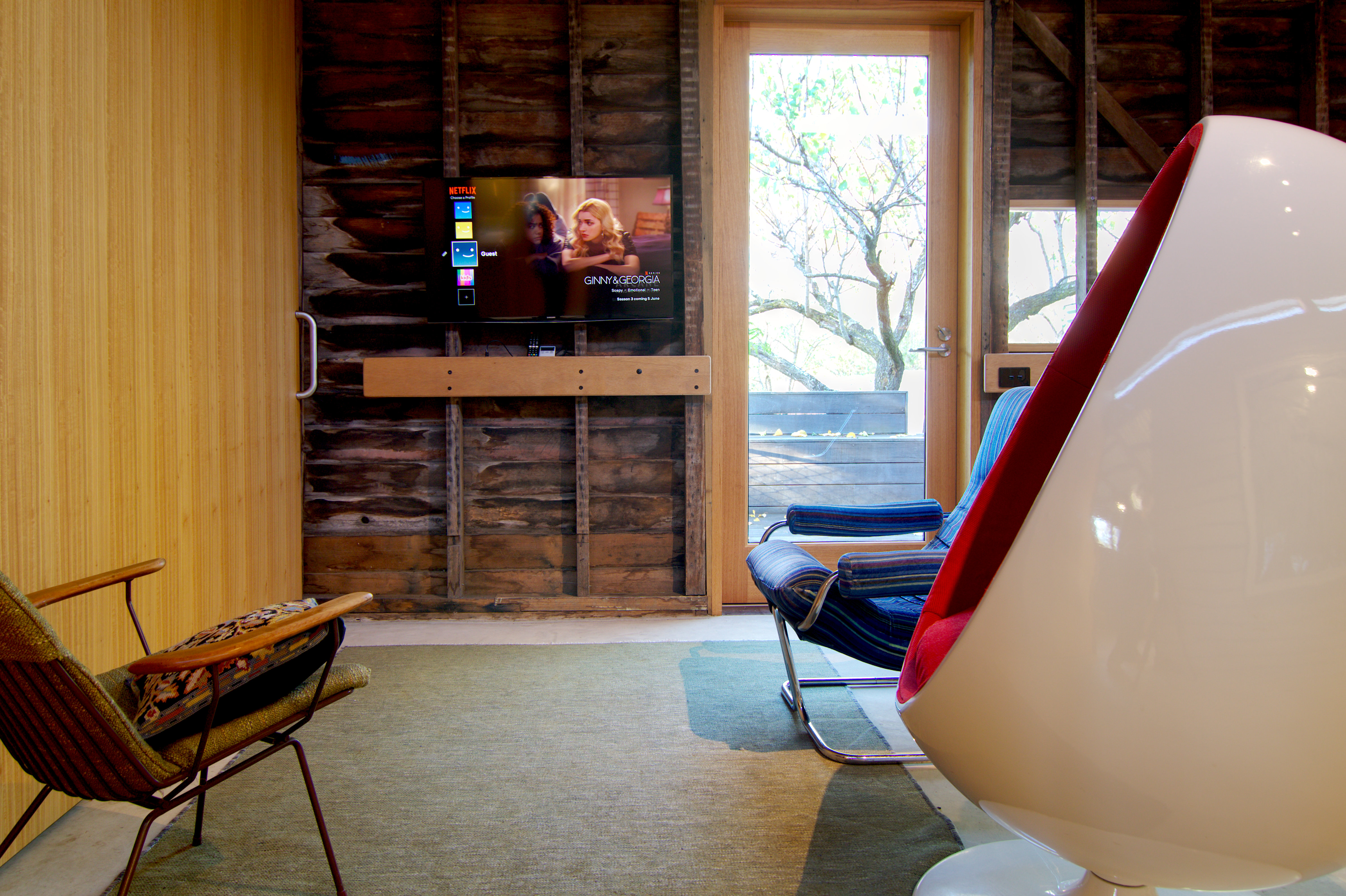 Living room with a wall mounted TV above a timber handrail and a glazed door out to a deck. Midcentury modern chairs grouped in the foreground.