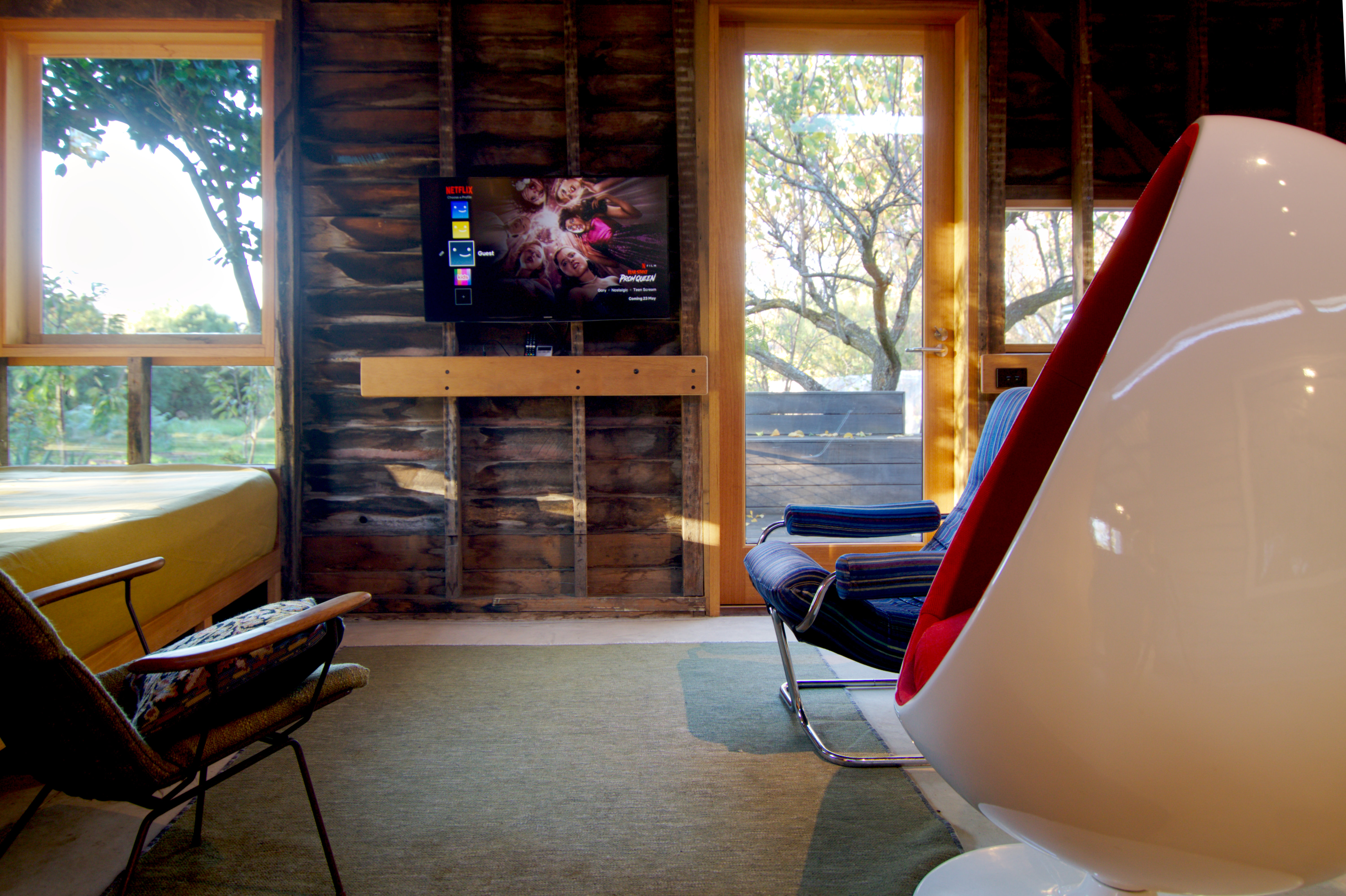 Ovalia egg chair with white shell and red upholstery in front of blue velhour chair to left. Metal chair and daybed in alcove to right. Timber wall in background with wall mounted TV above handrail and windows either side with view to deck and trees.