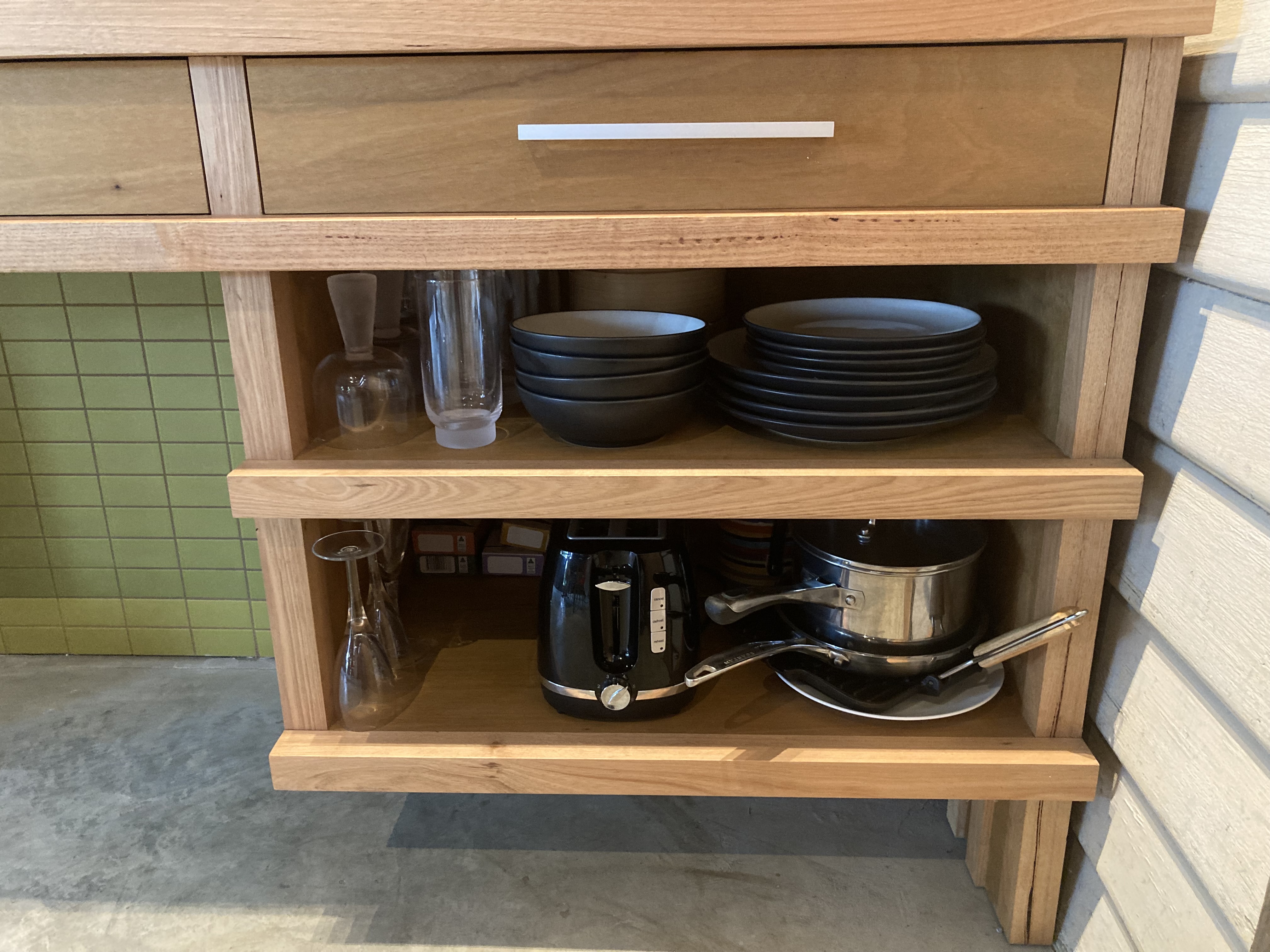 Timber drawer with shelves under containing glasses, plates and saucepans. Concrete floor and green mosaic tiled wall behind.