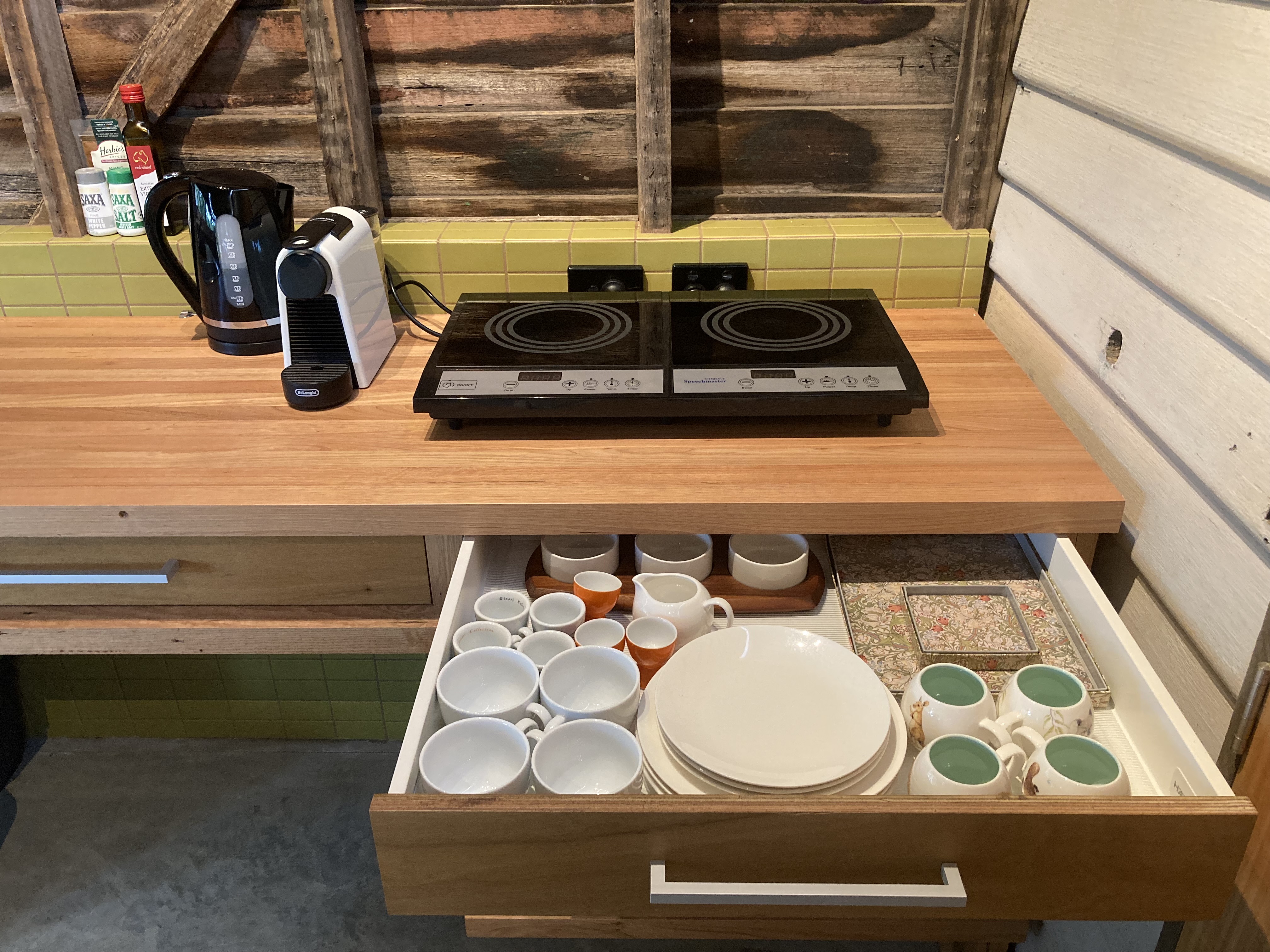 Closeup of kitchen bench with drawer open showing cups and plates. On bench, 2 burner cooktop, coffee pod machine & electric kettle. Green tiled splashback shelf and timber walls behind.