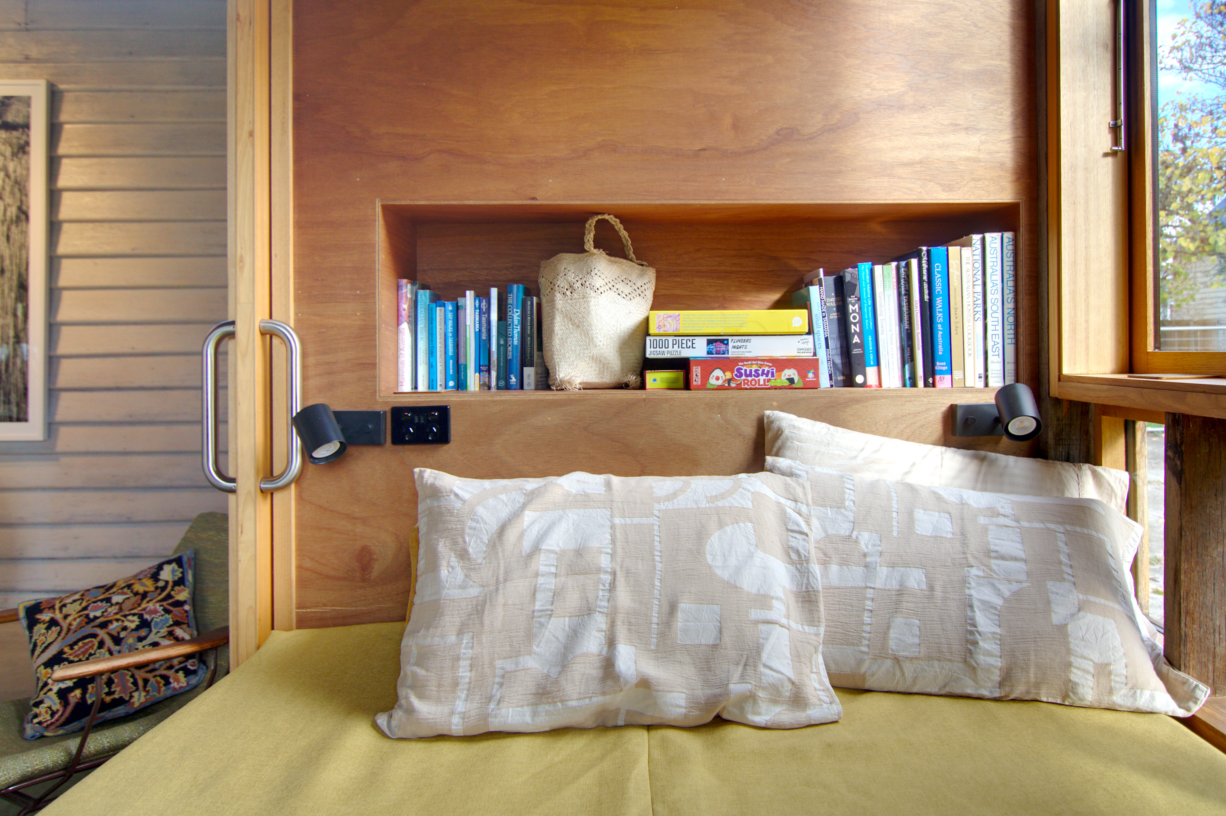 Plywood wall behind bed with inset shelf containing books and games. Sliding door handle to left and window to right.