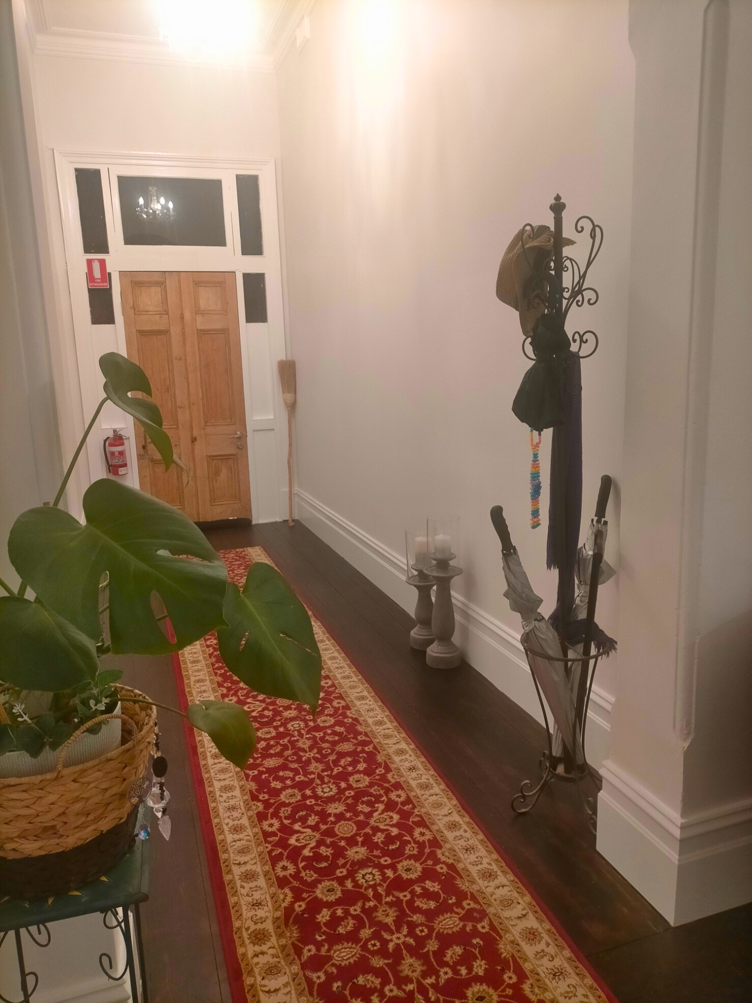 This image shows an indoor hallway with a red patterned runner rug and a dark wood floor. A coat rack with umbrellas and a hat is on the right, and a potted plant is on the left. A double door is visible at the end of the hallway. There are also decorative items like candles and a bessom