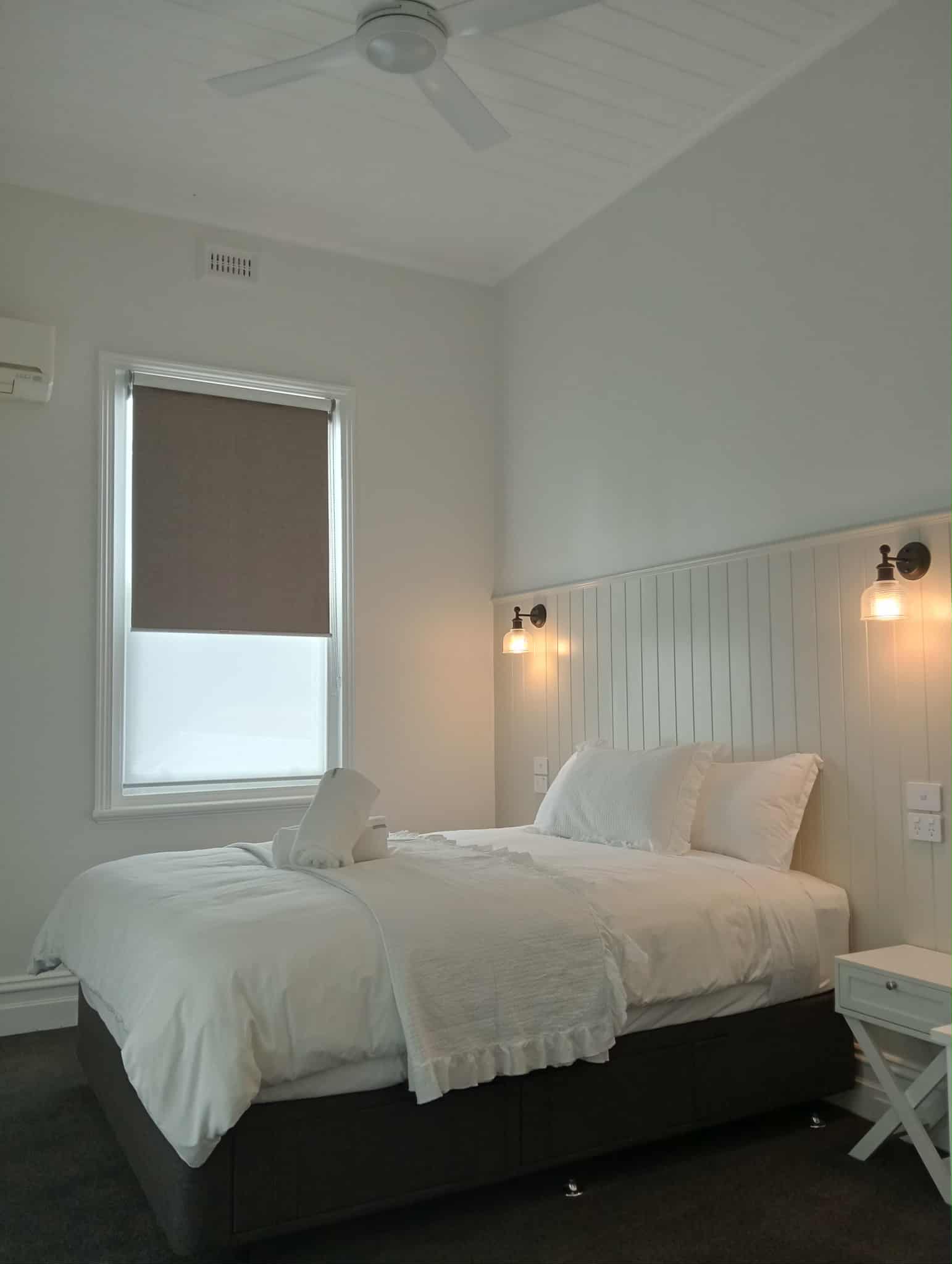 This image shows a bedroom with a king bed, nightstand, window, and ceiling fan. The walls are painted white, and there are two sconces on the wall above the headboard. The bed is made with white linens, and there are two towels folded on top. The window has a brown roller shade, and there is a small air conditioner on the wall to the left. The floor is dark carpet