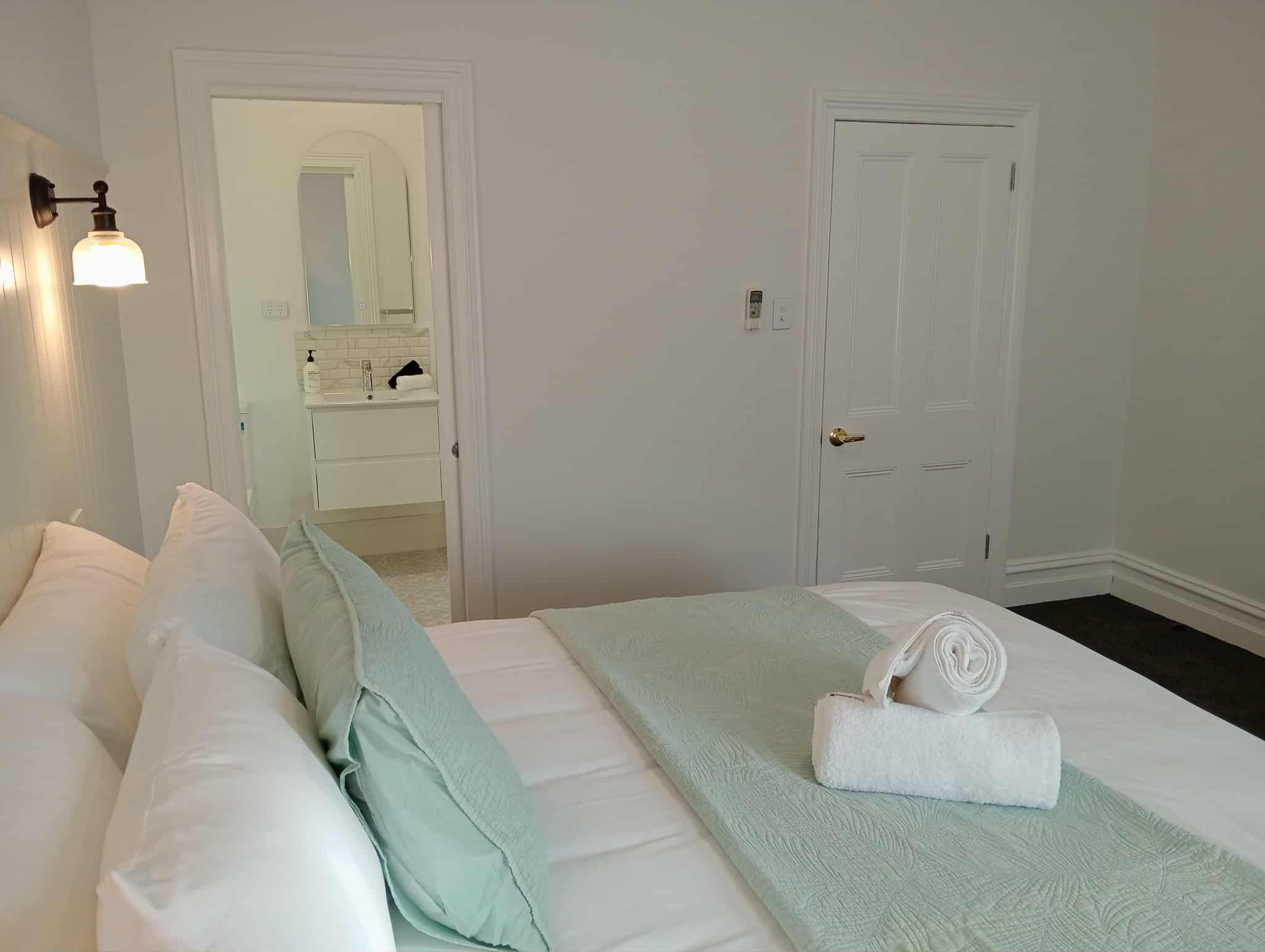 A bedroom with a King bed, white bedding, a light green throw blanket, and rolled-up white towels is shown. A doorway to a bathroom is visible, and a closed white door is to the right. A black wall sconce is on the left wall. The room has white walls and dark carpet