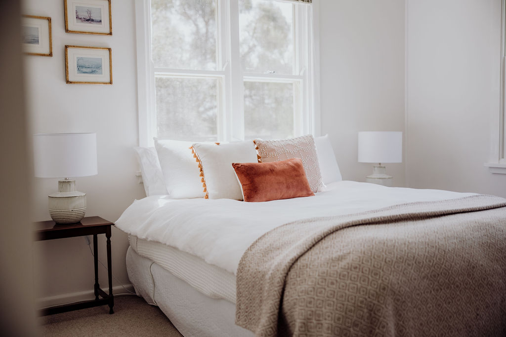 Bright and serene bedroom with a queen-sized bed, crisp white linens, and soft accent cushions. Large windows allow natural light to fill the space. Framed artwork and soft lighting create a calm and welcoming atmosphere.