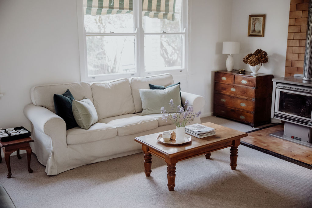 Light-filled lounge room featuring a cushioned sofa with pastel pillows, classic wooden furnishings, and a fireplace for warmth. Soft lighting, fresh flowers, and thoughtful styling create a relaxed, country charm.