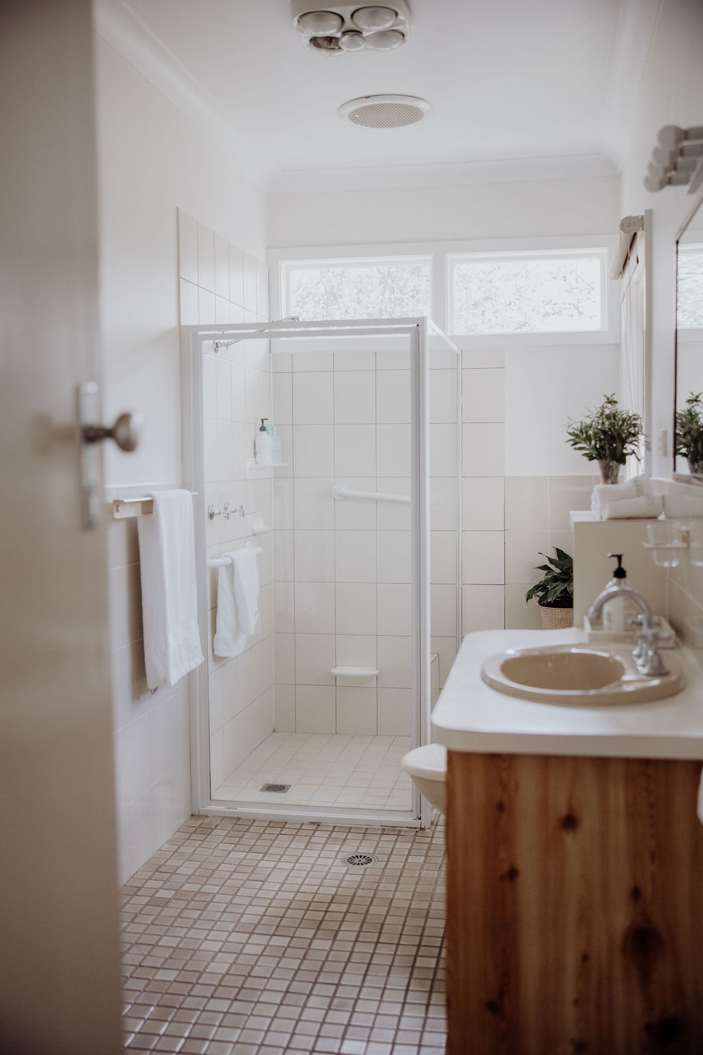 Bright, clean bathroom featuring a walk-in shower with glass doors and white tiled walls. The shower includes a horizontal grab rail for accessibility and built-in soap shelf. The vanity has a beige basin, and the space is decorated with neatly folded towels, offering both function and comfort.