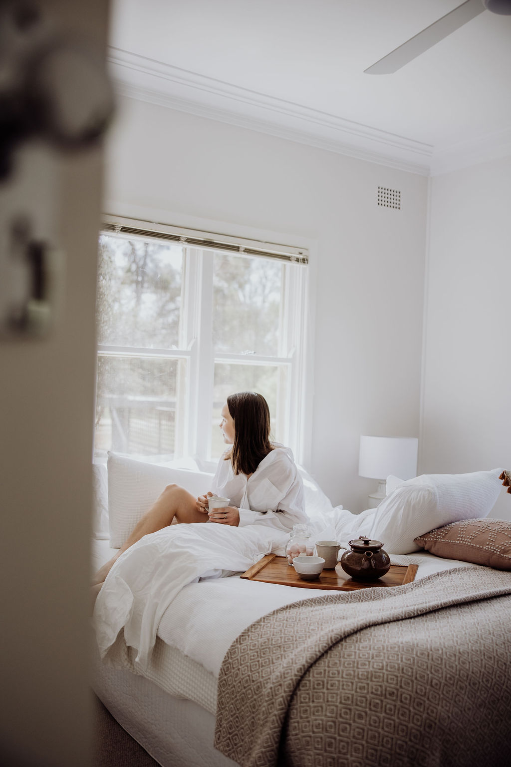 Peaceful bedroom with soft, neutral tones and cosy textures. A guest relaxes with a cup of tea on the bed, surrounded by calming decor and natural light streaming through large windows.