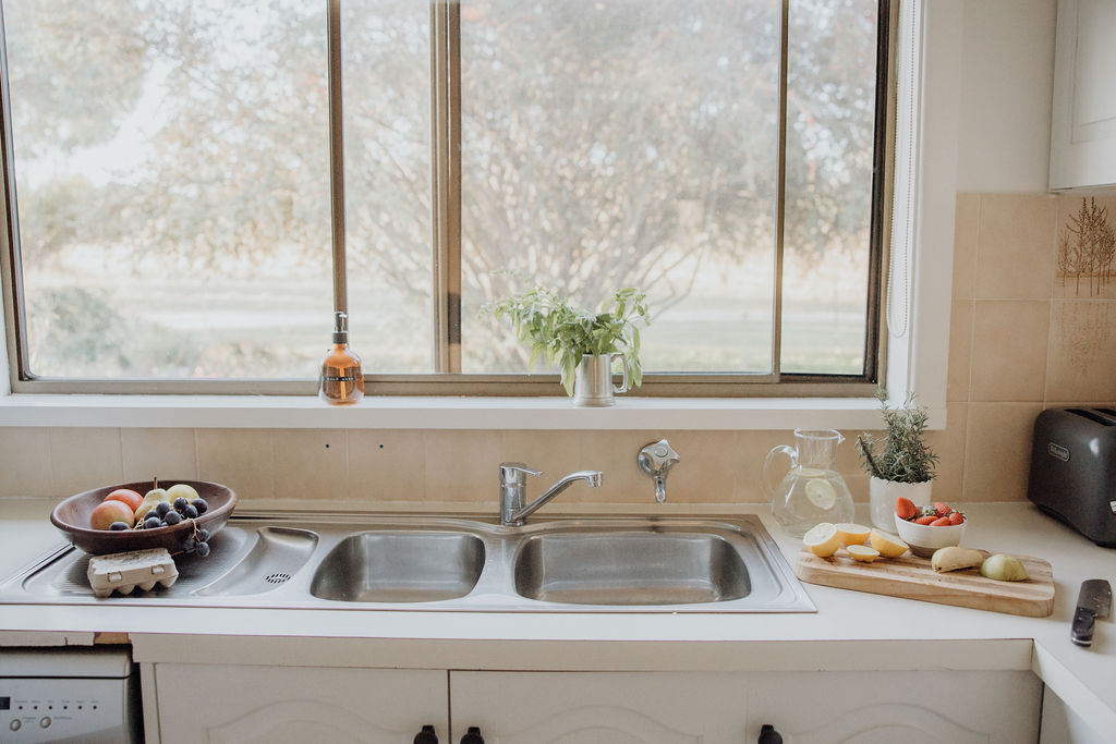 Sunlit kitchen sink area with a large window overlooking the garden. A double stainless steel sink is flanked by a bowl of fresh fruit and eggs on the left, and a wooden chopping board on the right holding sliced lemons, ginger, and a bowl of cherry tomatoes. Fresh herbs, a glass jug of lemon water, and a toaster add a homely, welcoming touch.
