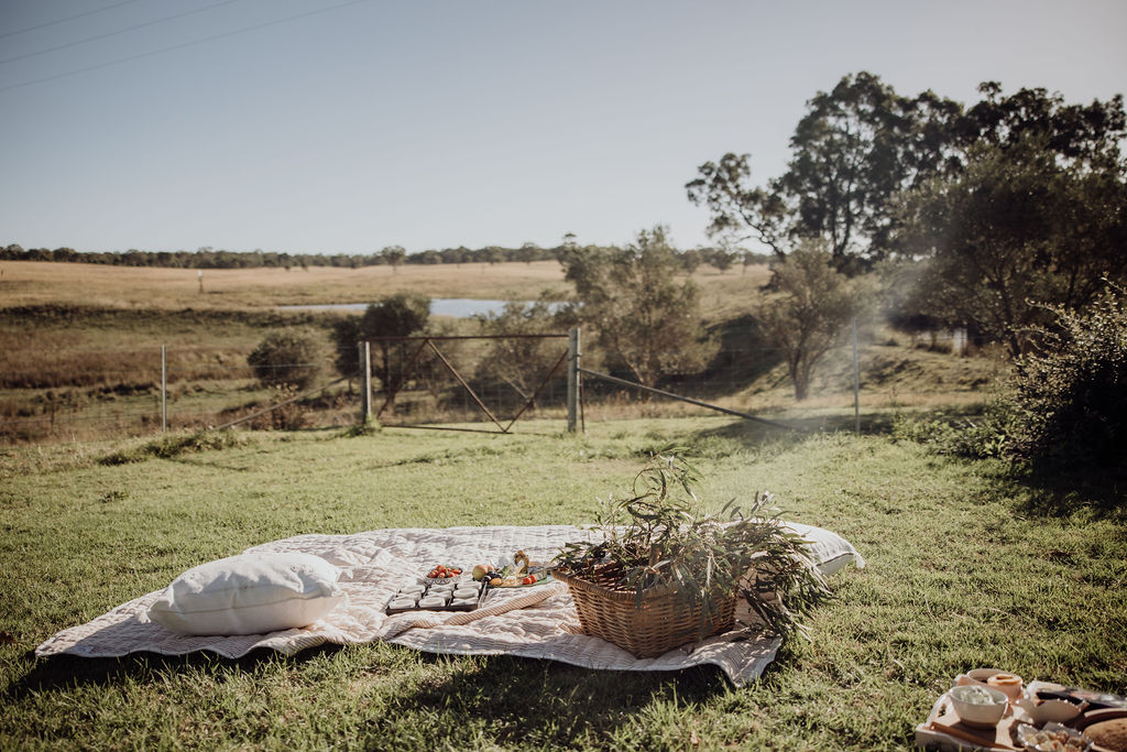A welcoming picnic blanket and basket of produce and foliage is set in a country garden with views across paddocks and a distant dam.n